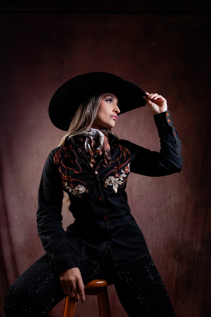 Stylish woman in western attire and cowboy hat on a stool, exuding confidence.