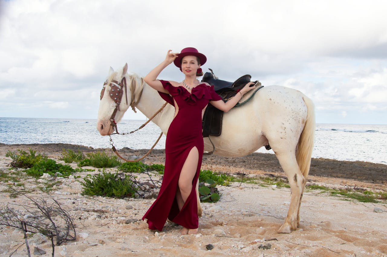A stylish woman in a red dress poses with a white horse on a scenic seaside beach.