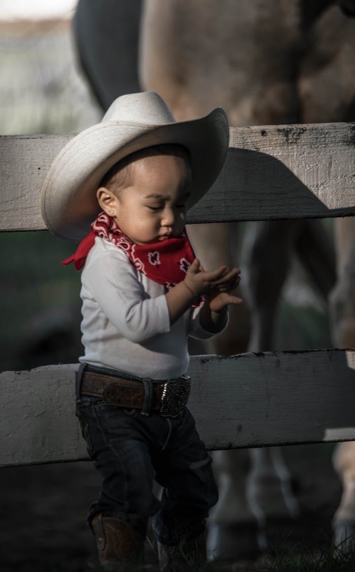 Adorable child dressed as a cowboy with hat and boots leaning against a fence.